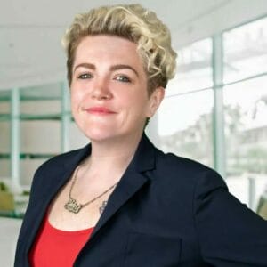 Professional woman with short curly hair, smiling, wearing a navy blazer and red top in a bright, modern office setting.