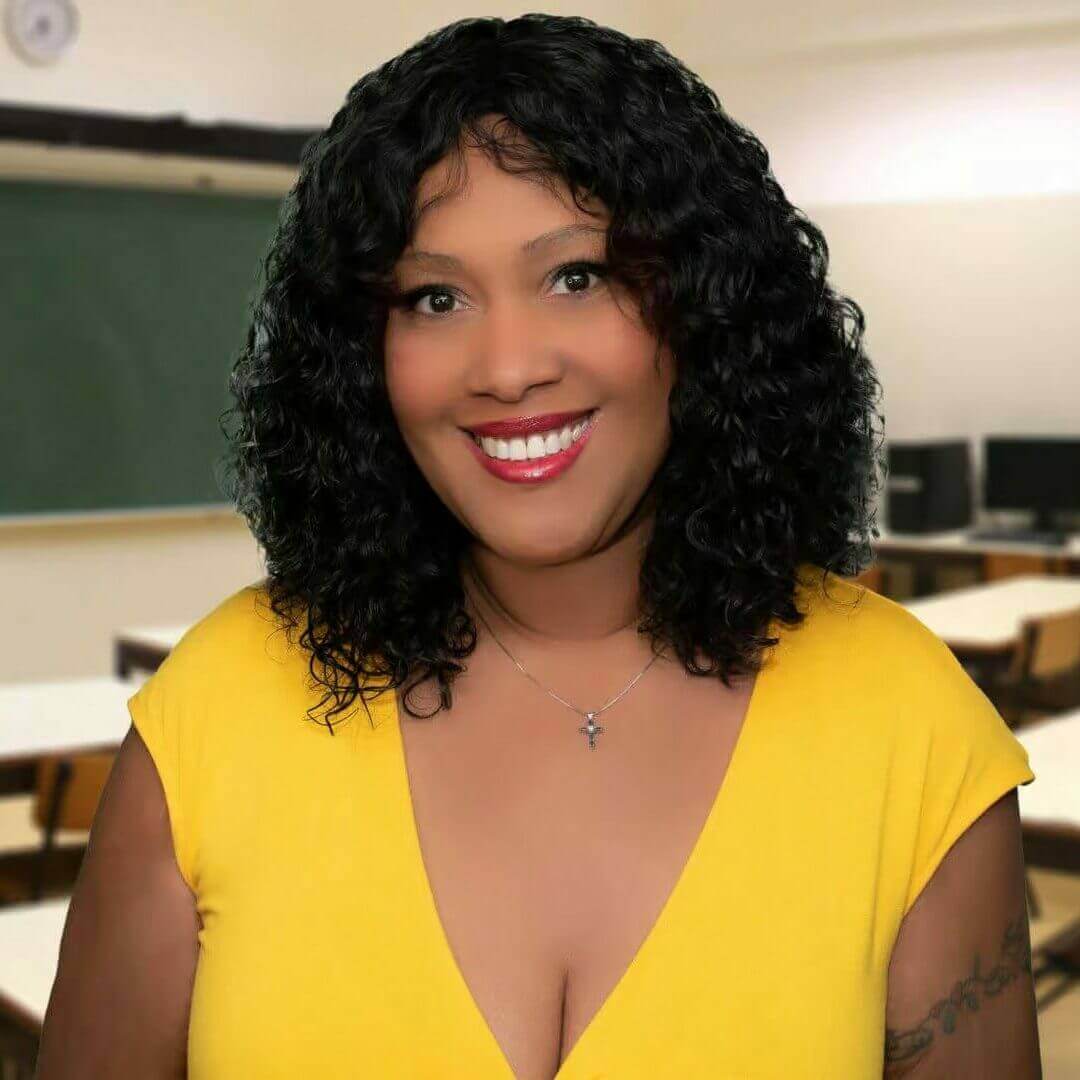 Smiling woman with curly hair in yellow dress, standing in a classroom with computer desks and blackboard in the background.