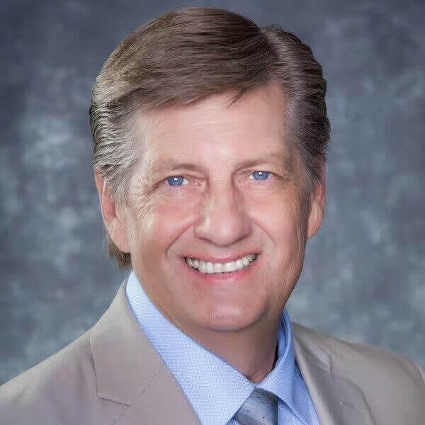 Smiling man in a beige suit and blue shirt against a gray background.