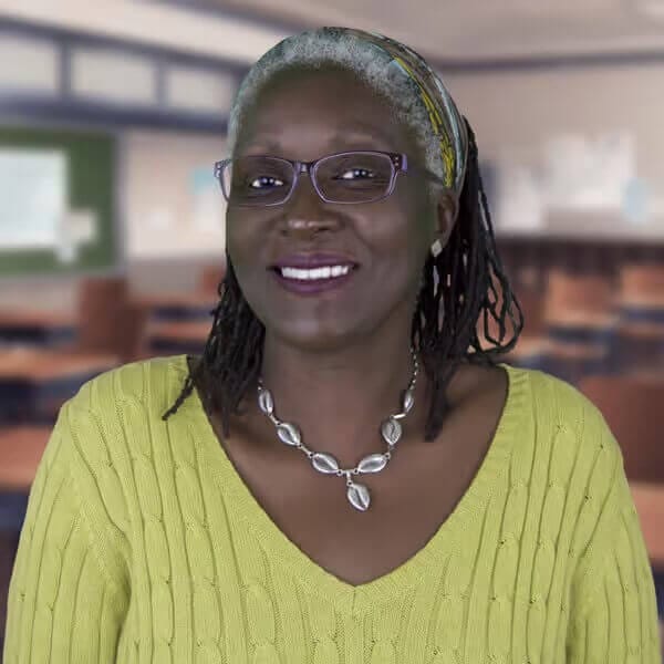 Smiling woman in a yellow sweater and glasses, standing in a classroom setting, wearing a necklace and headband.