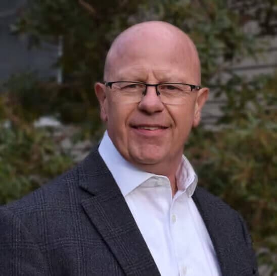 Bald man in glasses wearing a suit, smiling outdoors with greenery background. Ideal for business profile or professional headshot.