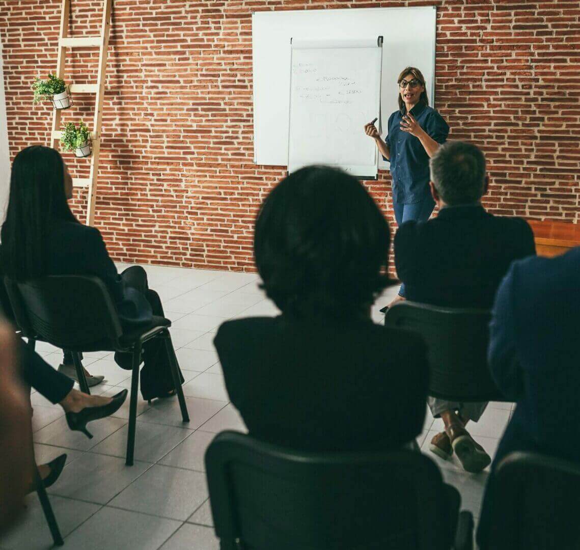 Woman coach explaining new business project to team in meeting room with brick wall background.