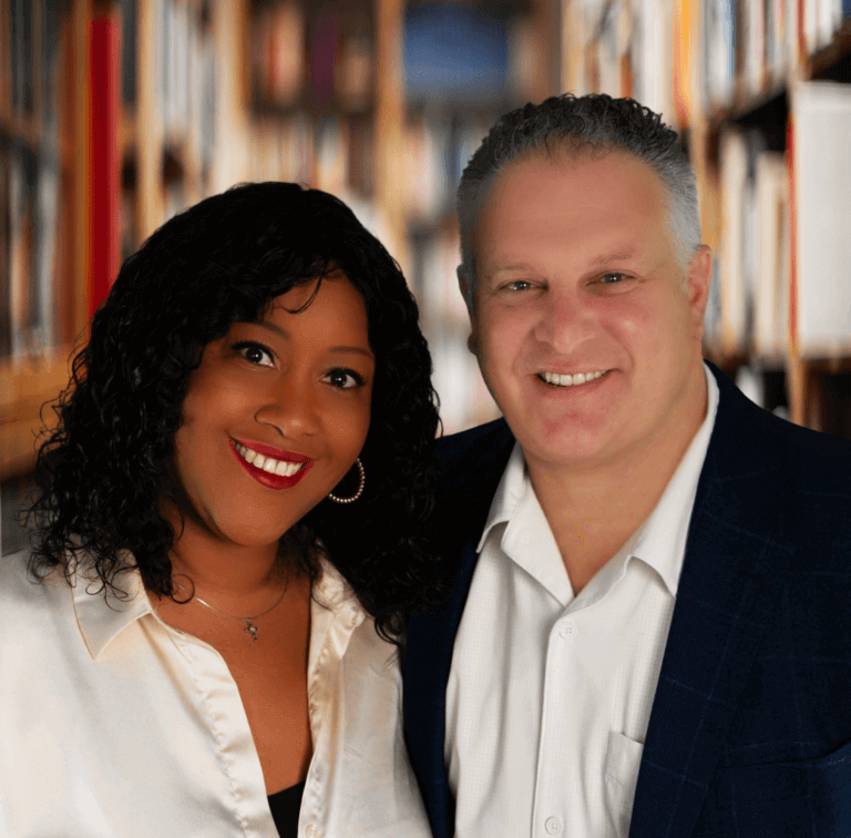 Couple smiling in a library setting, diverse duo, professional attire, books in background, friendly atmosphere.